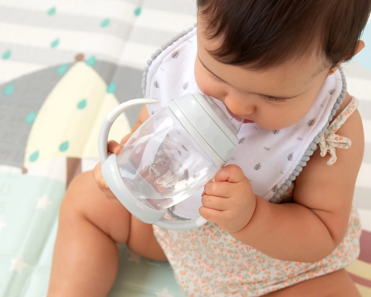 Bebé a beber de um copo infantil transparente com pega dupla e tampa branca