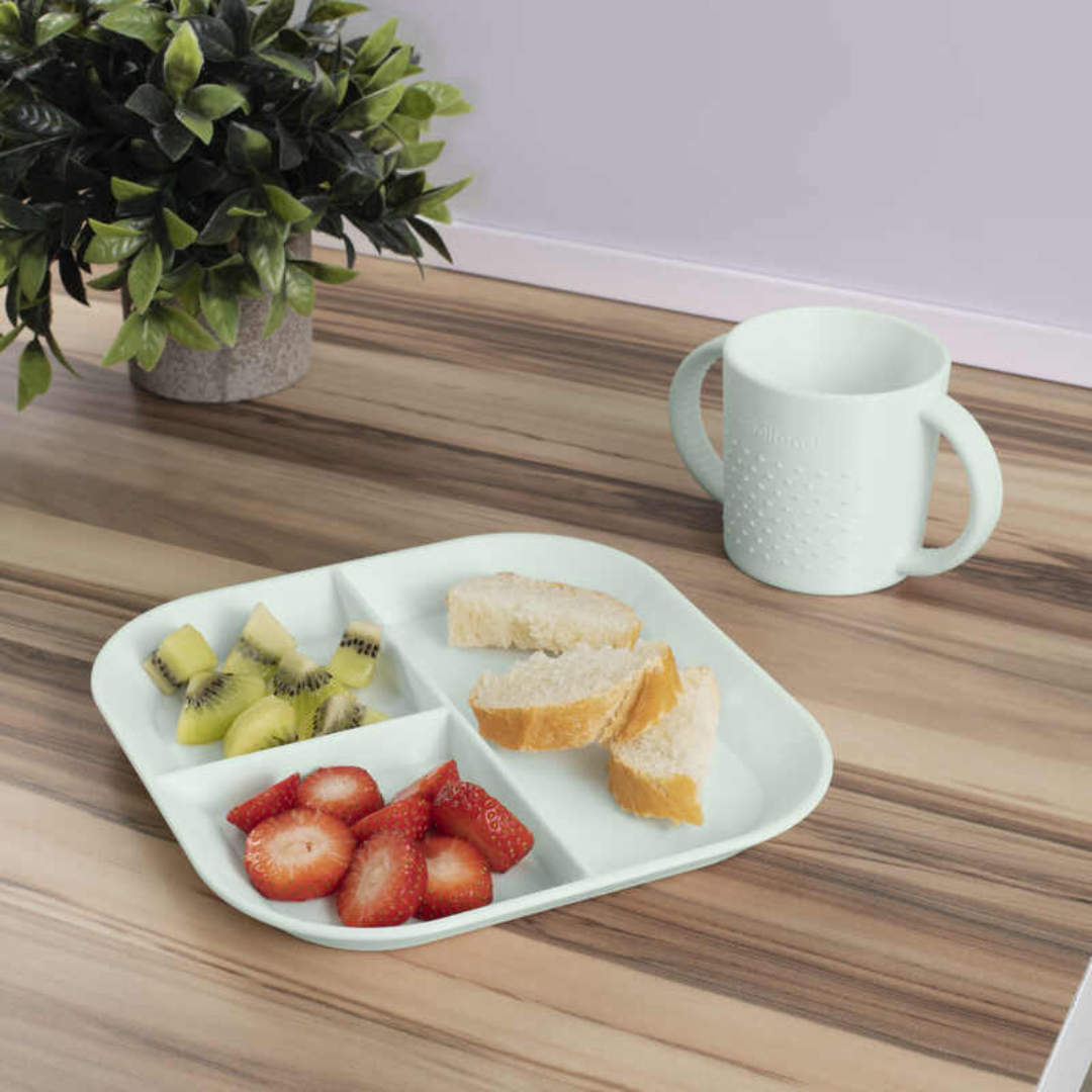 Prato dividido branco com frutas e pão, copo branco com duas pegas, mesa de madeira e planta artificial