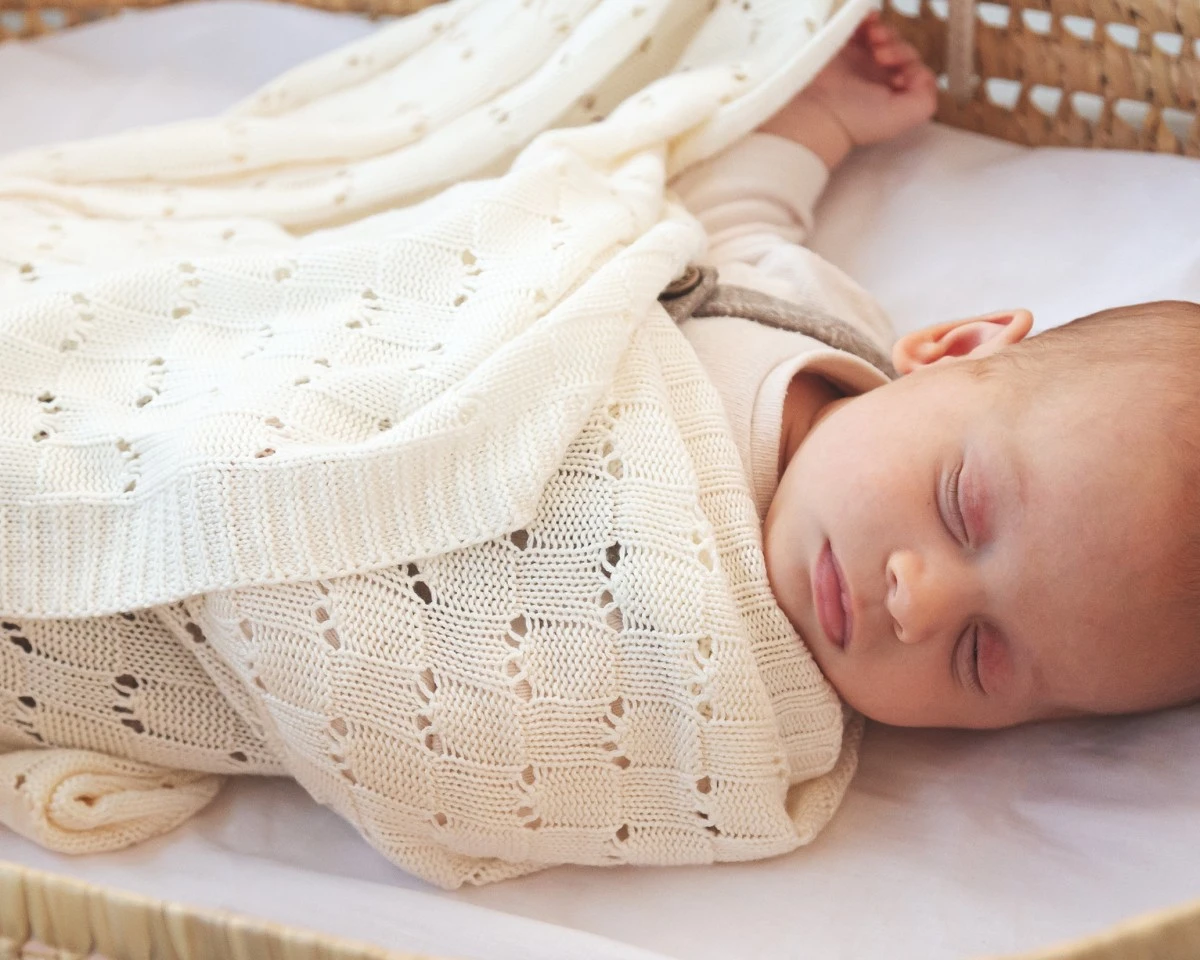 Bebé dormindo envolto em manta de malha creme num berço claro