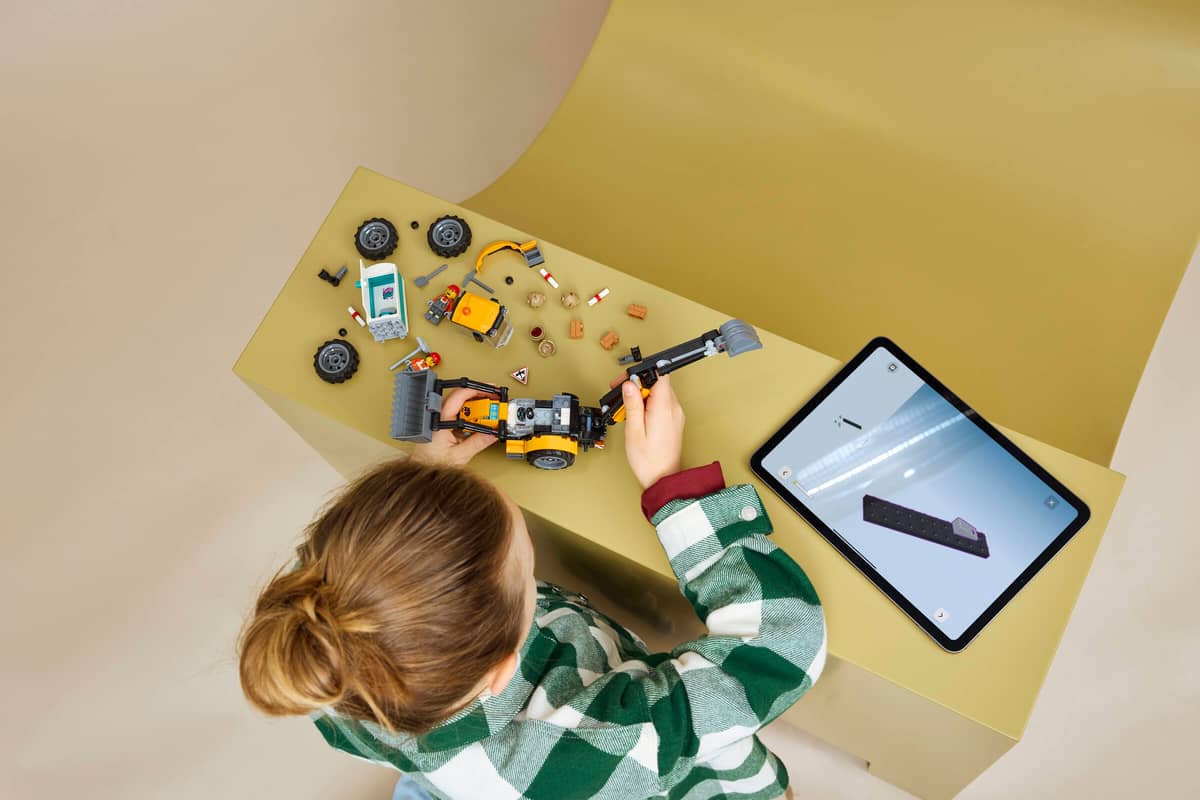 Criança a montar brinquedo de construção amarelo com escavadora junto a tablet com instruções