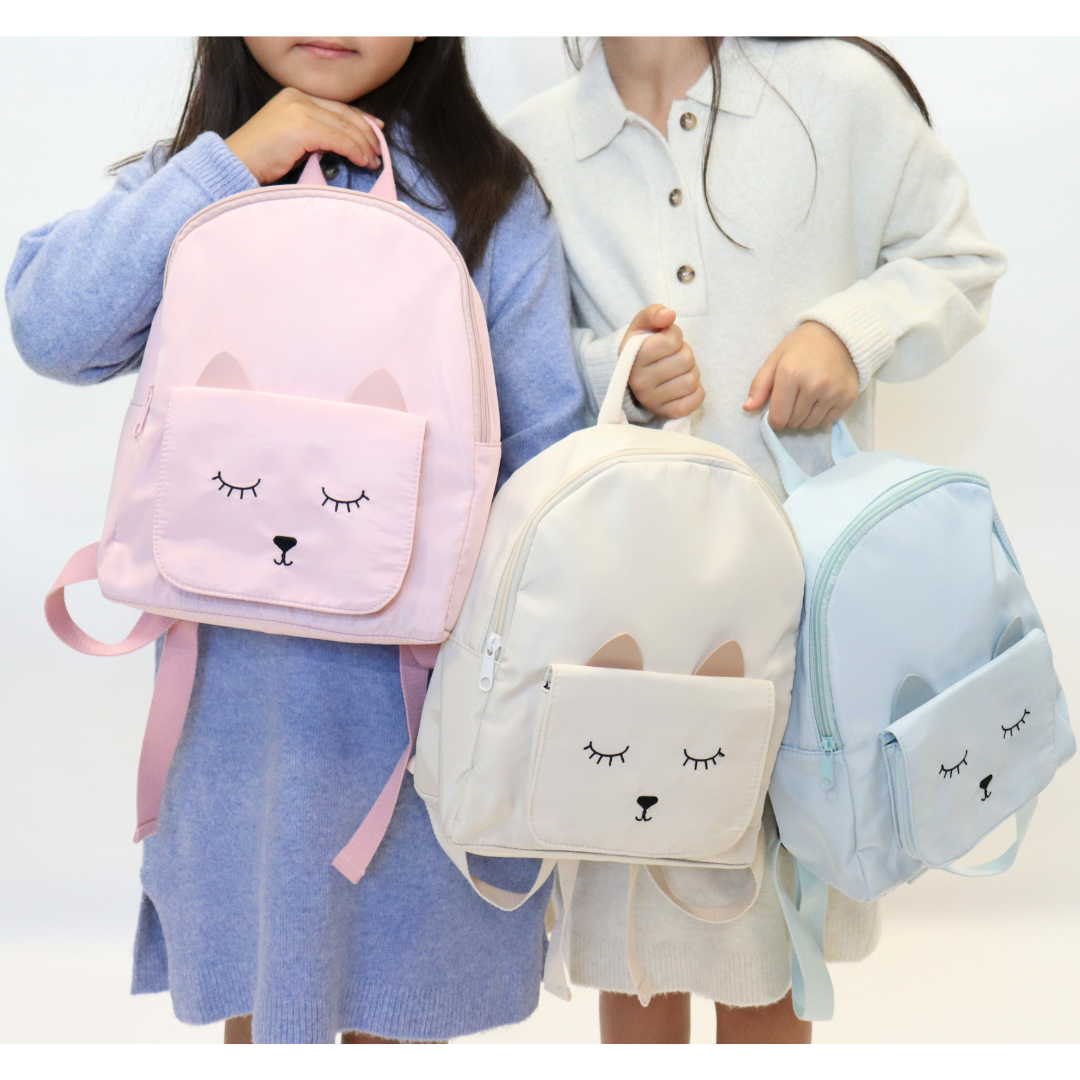 Mochilas infantis rosa, branca e azul claro com rosto de animal desenhado e orelhas, seguradas por crianças.