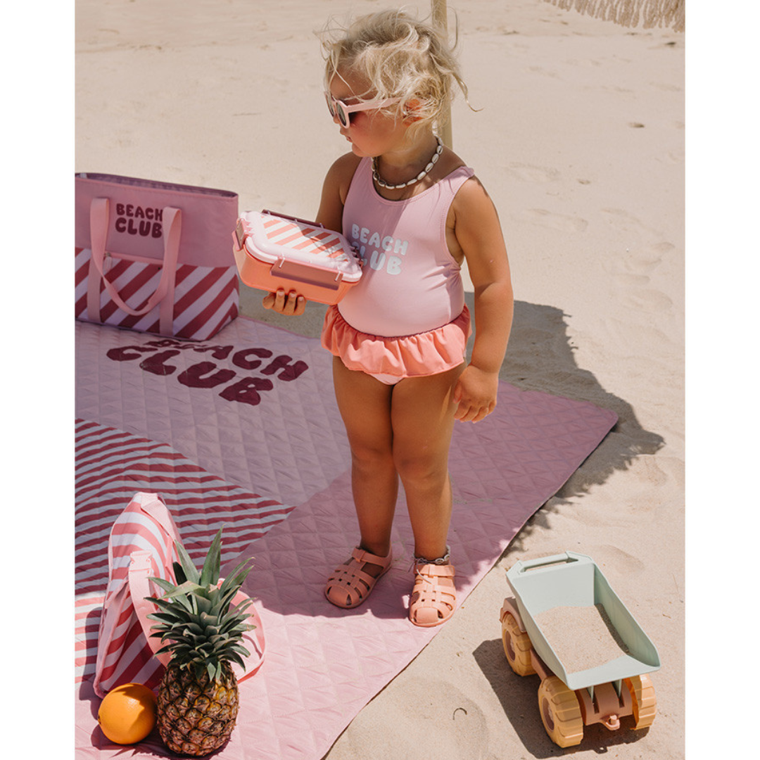 Menina com fato de banho rosa BEATCH CLUB na praia com toalha rosa, saco, ananás e brinquedo camião de areia