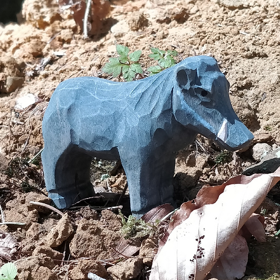 Pequena estátua de javali esculpida em madeira cinza-azulada no chão de terra