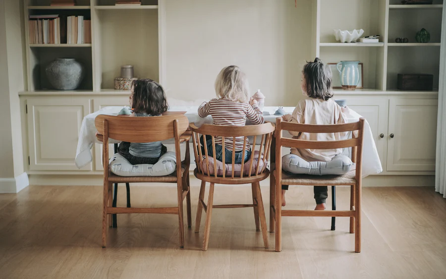 Três crianças sentadas em cadeiras de madeira clara com almofadas cinzentas numa sala interior