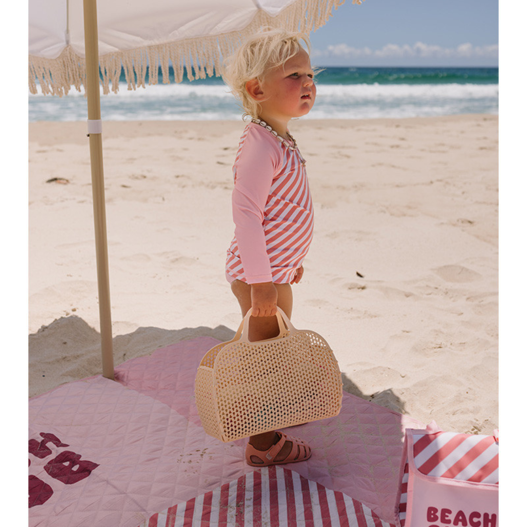Criança na praia com fato de banho rosa às riscas e bolsa bege de plástico perfurado