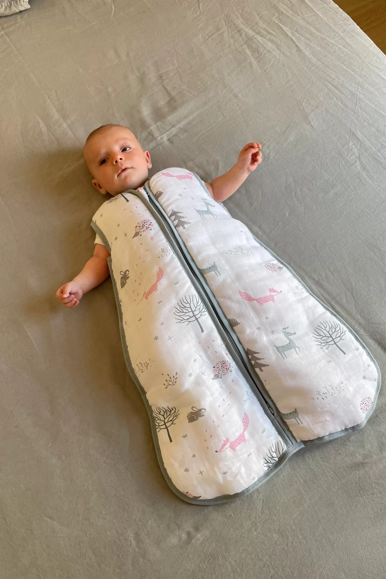 Saco de dormir para bebé branco com padrão animal e árvores em cama cinza.