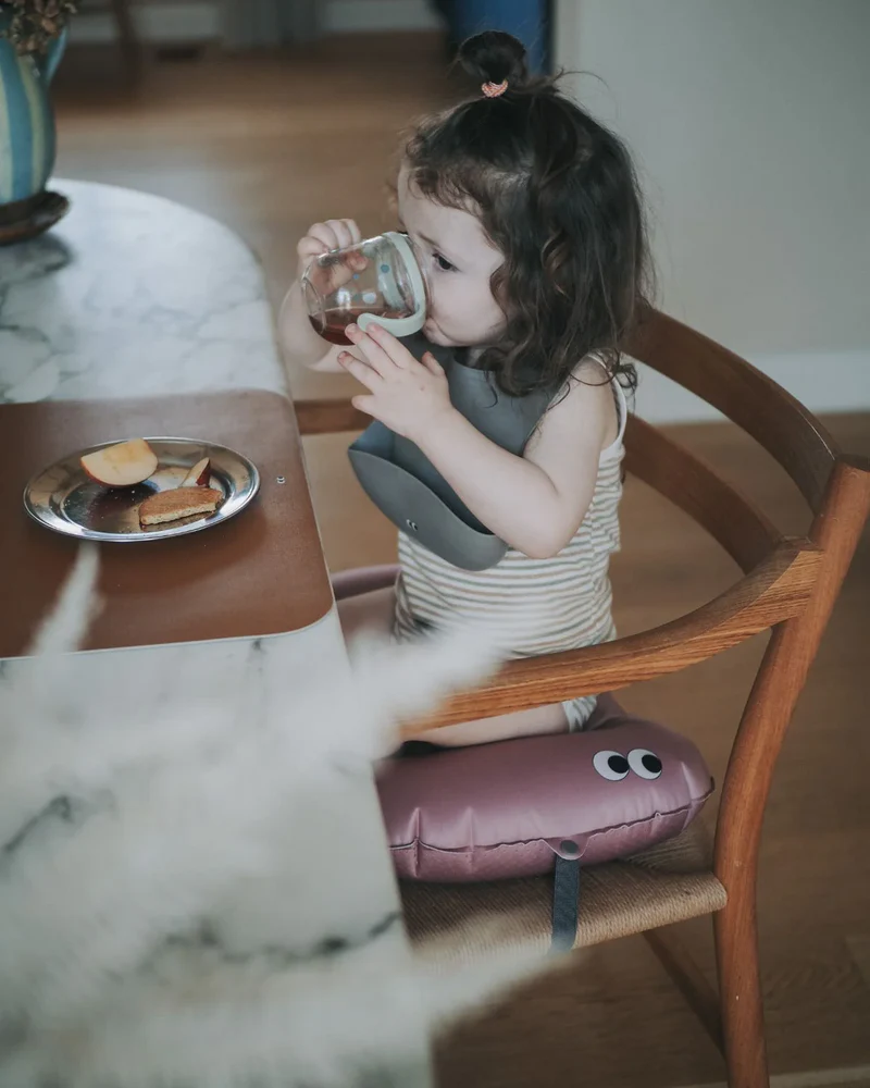 Criança sentada numa cadeira com babete a beber de copo, almofada roxa com olhos sob a cadeira e prato com maçã e bolachas na mesa
