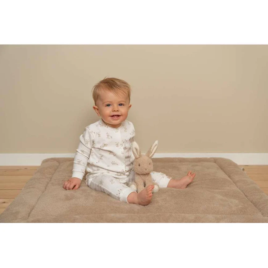 Bebé com pijama branco e coelho de peluche sentado num colchão felpudo bege