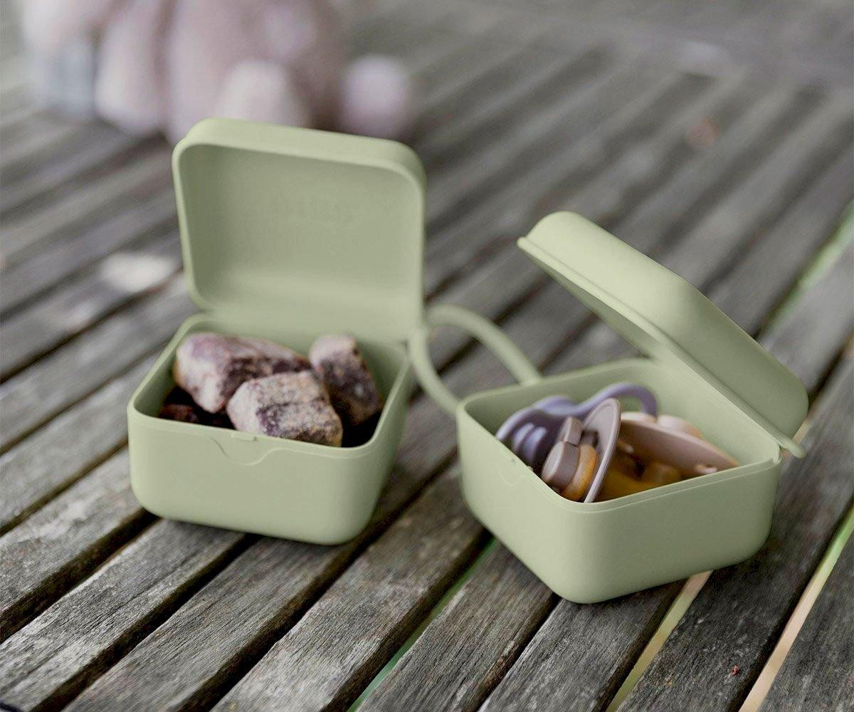 Duas caixas verdes pequenas abertas contendo pedras e chupetas, sobre mesa de madeira cinzenta.