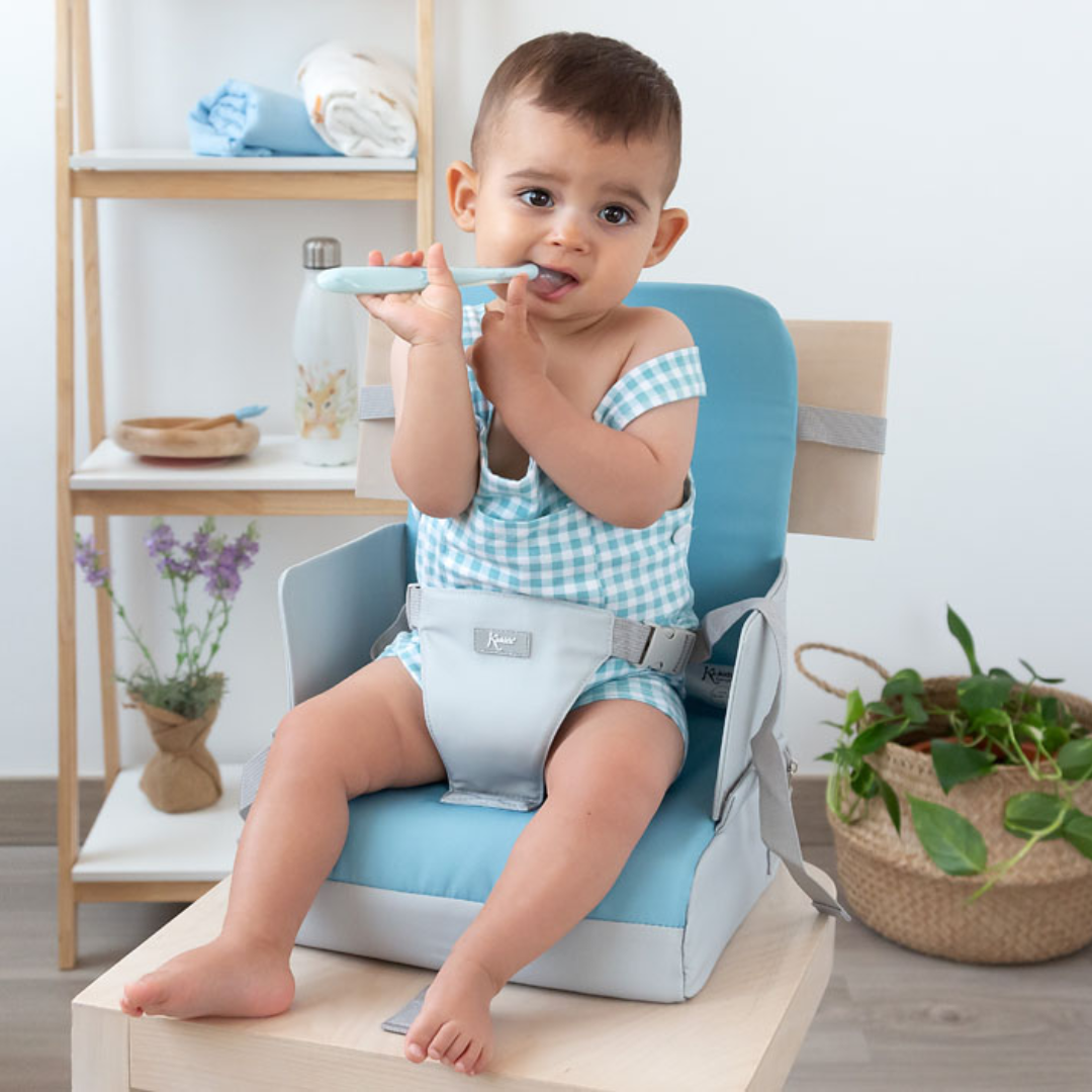 Bebé sentado numa cadeira de refeição portátil azul claro com cinto cinza a segurar uma colher azul e branca