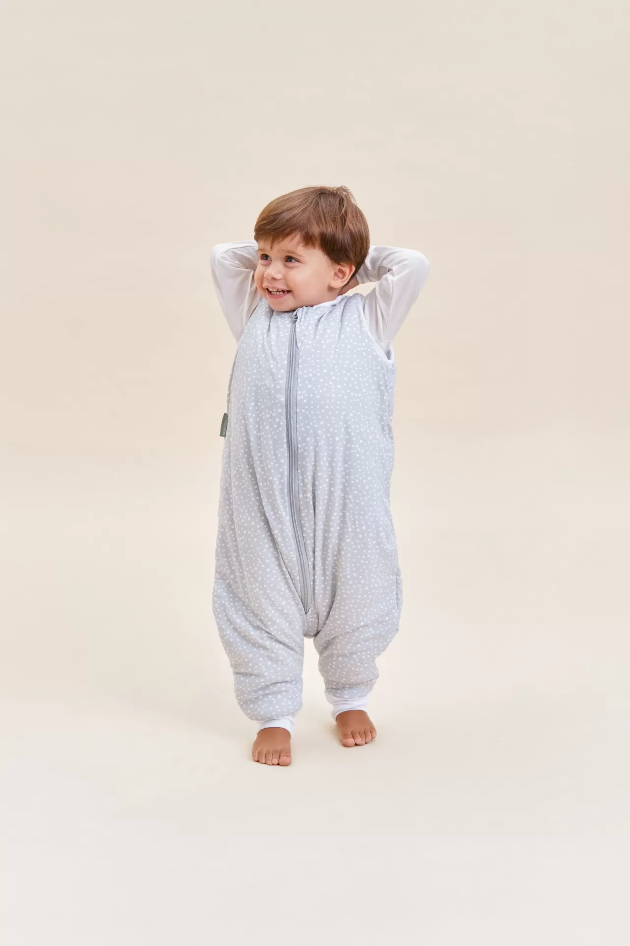 Bebé com macacão cinzento claro com padrão de pontos brancos descalço num fundo bege claro