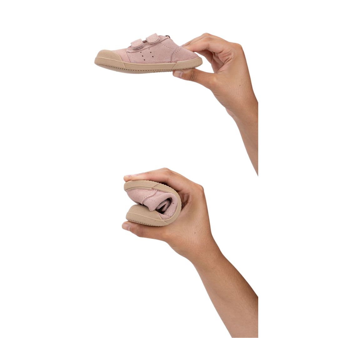Sapato infantil rosa com sola beige flexível e duas tiras de velcro, segurado por mãos, imagem mostra sapato estendido e dobrado.