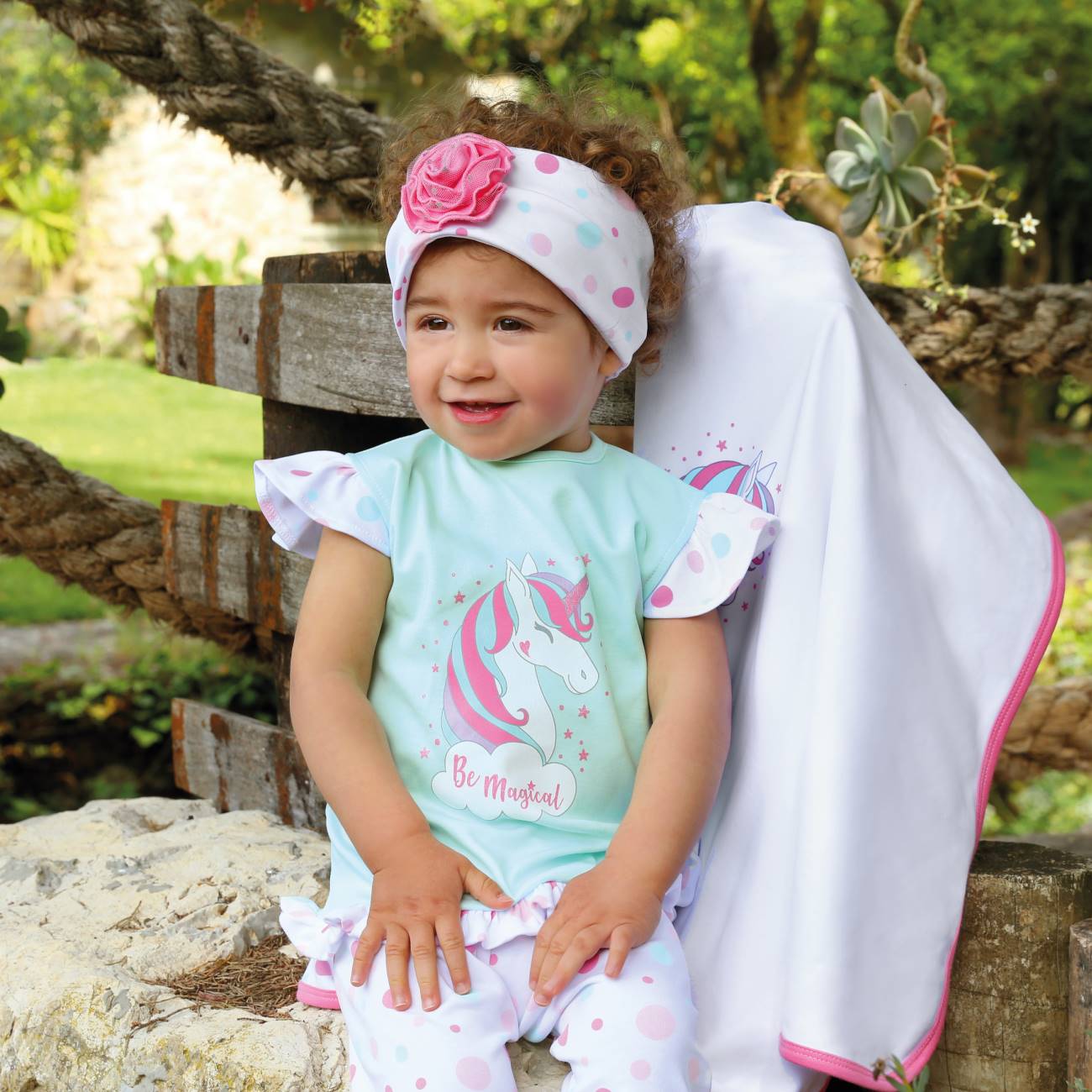 Bebé vestindo roupa com unicórnio e faixa com flor rosa, sentado ao ar livre com cobertor branco com borda rosa