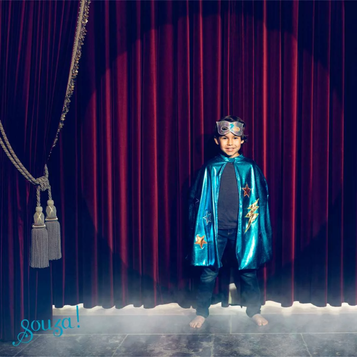 Criança em palco com capa azul e máscara, cortina vermelha ao fundo