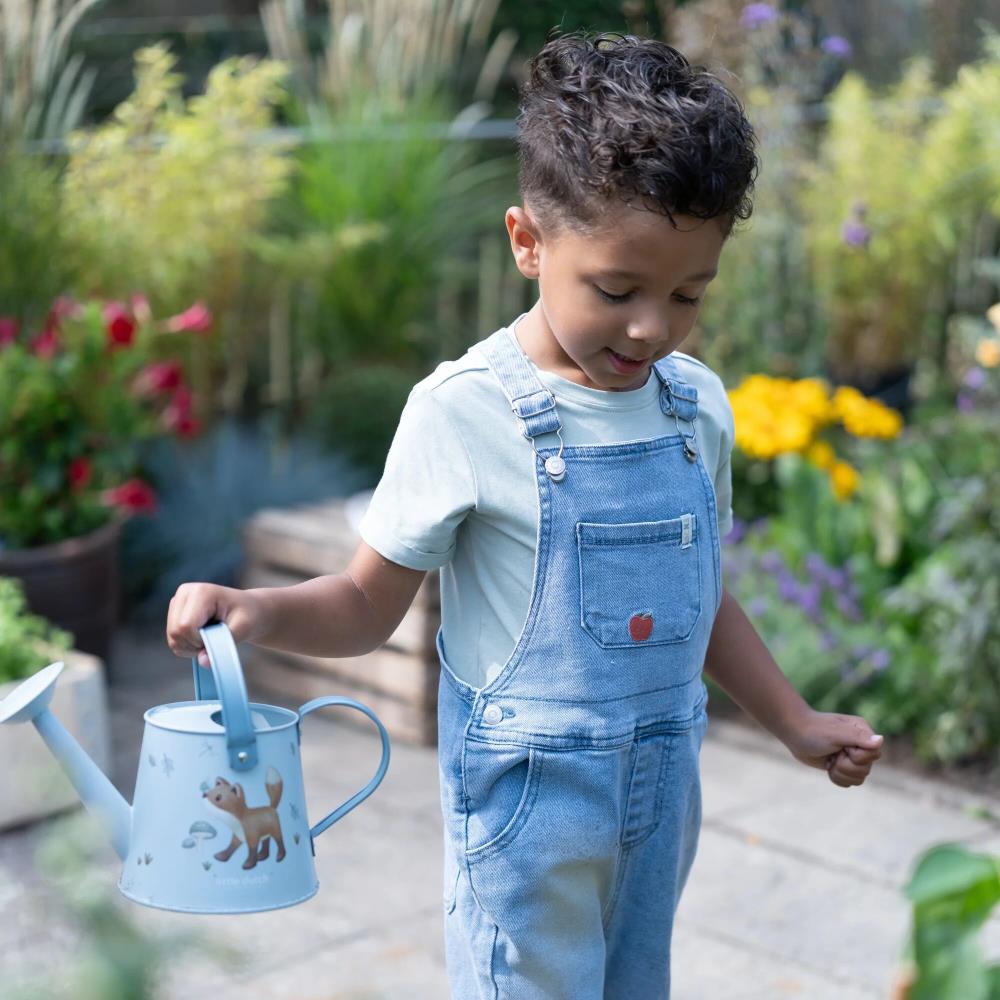 Criança com jardineiras azuis segurando regador azul claro num jardim