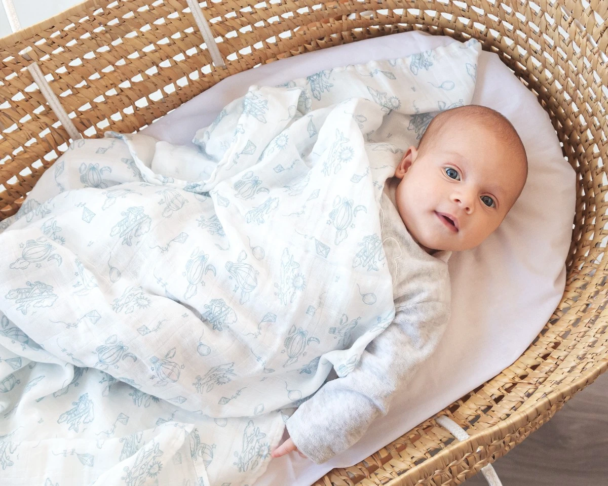 Bebé deitado num berço de verga coberto por uma manta branca com estampado azul claro.