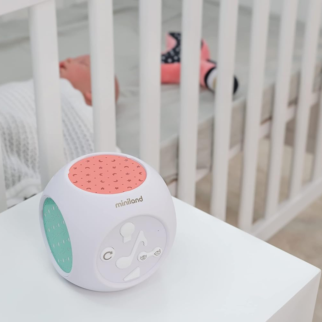 Cubo musical branco com detalhes verdes e cor-de-rosa com estrelas, junto a berço com bebé a dormir