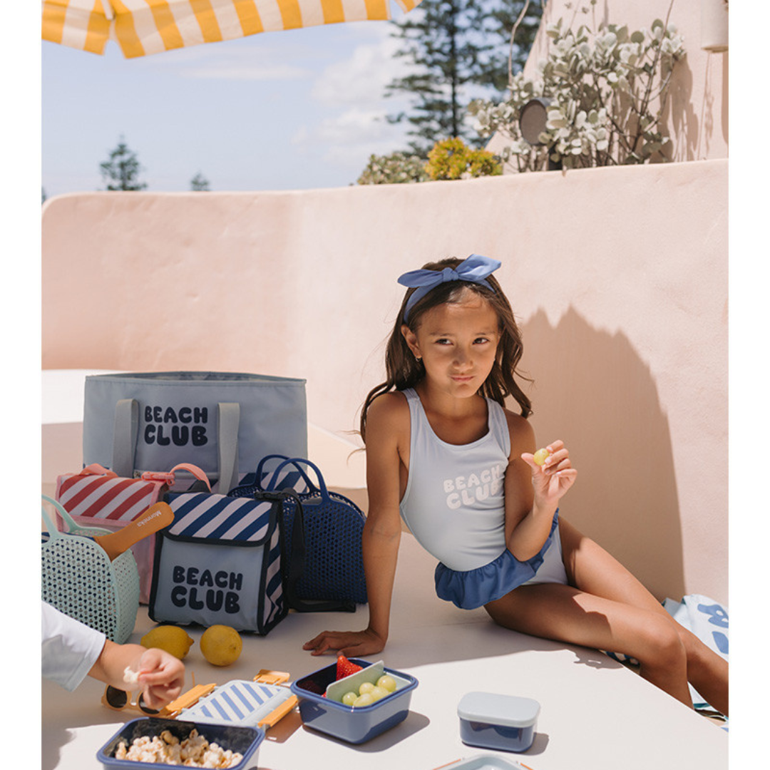 Criança com fato de banho azul claro e faixa azul com sacos BEACH CLUB azuis sobre mesa branca