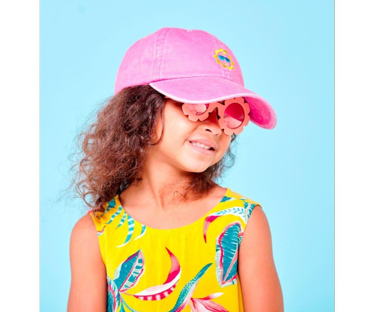 Criança com vestido amarelo e boné rosa e óculos de sol florais frente a fundo azul claro