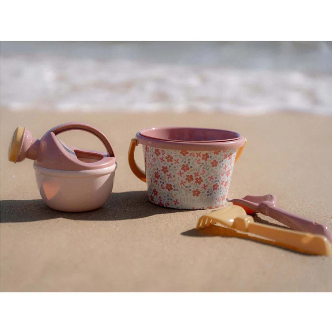Conjunto de brinquedos de praia em plástico com regador, balde floral e pás sobre areia próximo ao mar