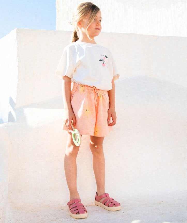 Menina vestida com t-shirt branca e calções cor de pêssego com sandálias rosas em fundo branco