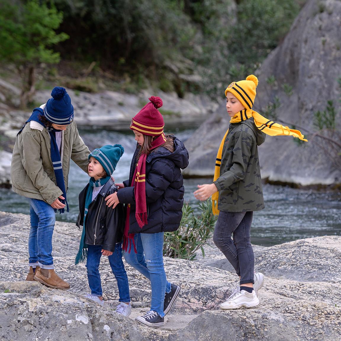 Quatro crianças com roupas de inverno coloridas junto a um rio rodeado de rochas.