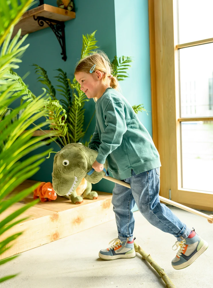 Brinquedo cavalo de pau dinossauro verde com criança numa sala com plantas