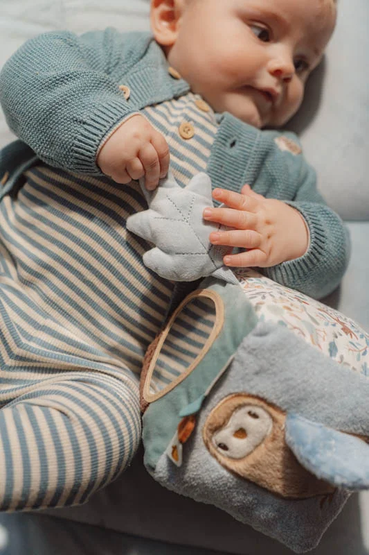 Bebé com fato de corpo às riscas e casaco verde segurando brinquedo de peluche com coruja