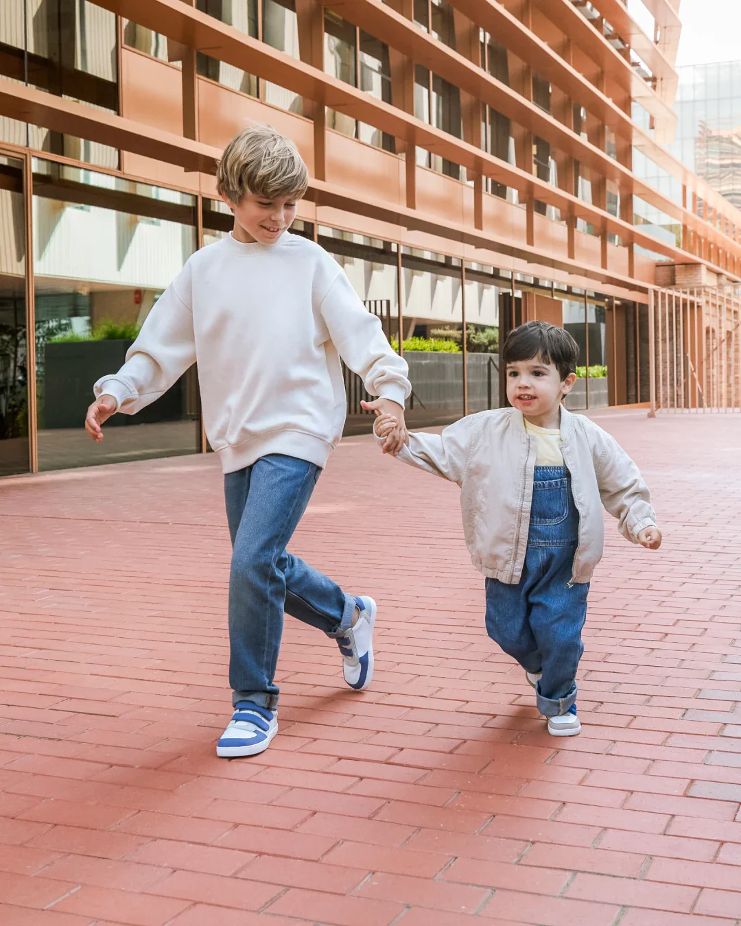 Duas crianças a andar de mão dada numa rua urbana com chão de tijolos vermelhos e edifício contemporâneo ao fundo.