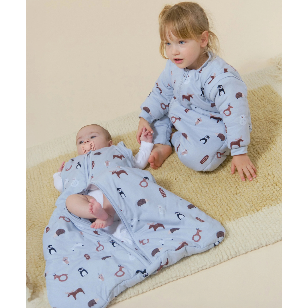 Dois bebés com roupas de dormir azuis estampadas sentados/ deitados sobre manta bege