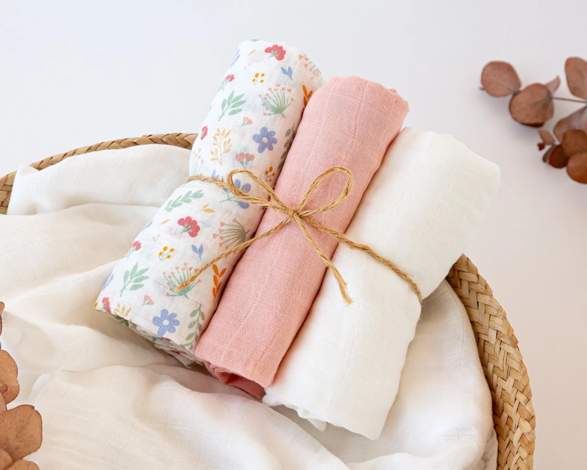 Cesta de vime com três toalhas enroladas de algodão ou linho, cores branco, rosa e floral, fundo branco minimalista
