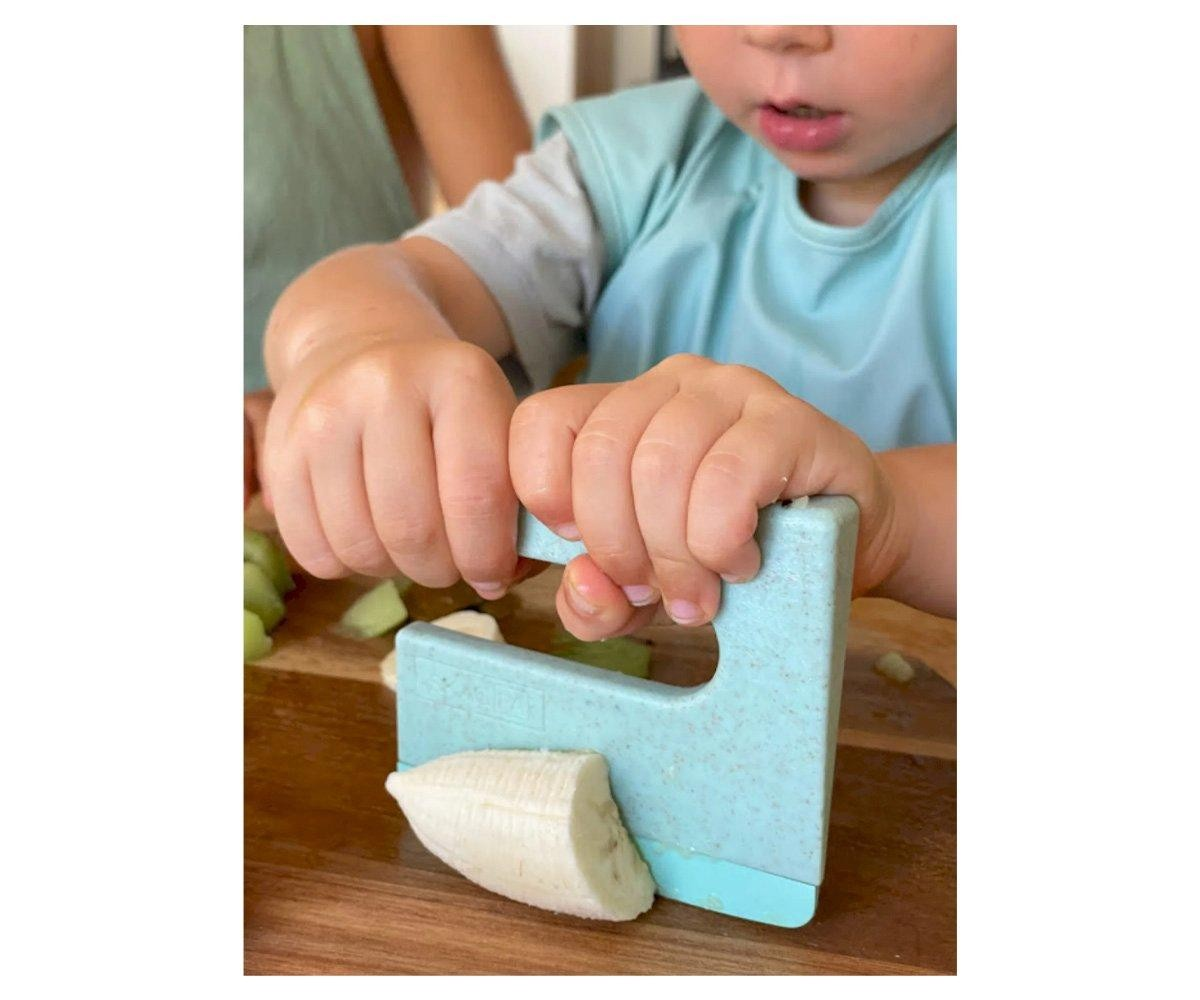 Criança a cortar uma banana com um cortador azul claro numa tábua de madeira.