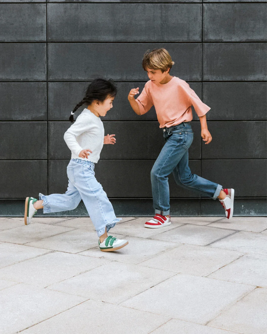 Duas crianças a correr vestindo roupas casuais e sapatilhas coloridas junto a um muro cinza.