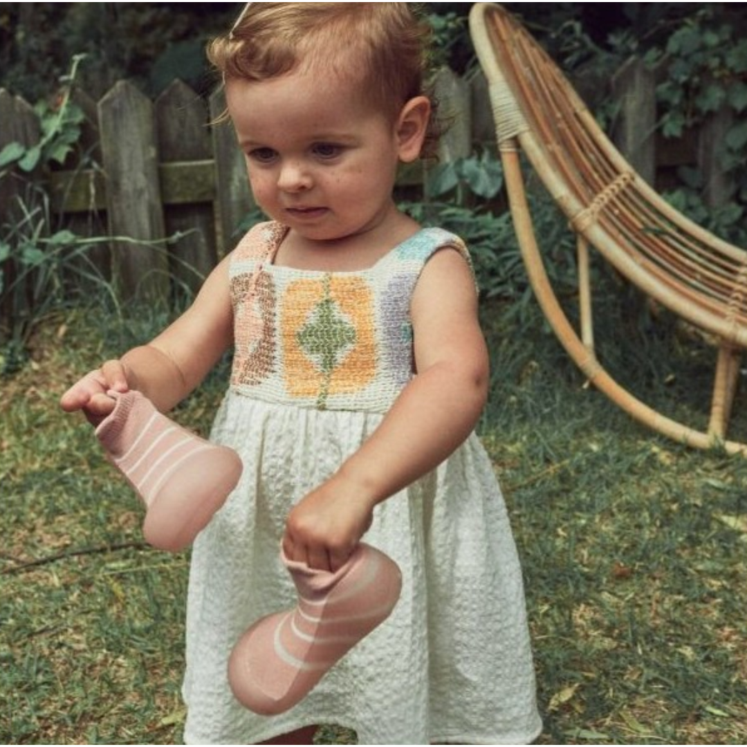 Criança com vestido branco e padrão colorido segurando meias cor-de-rosa com riscas num jardim