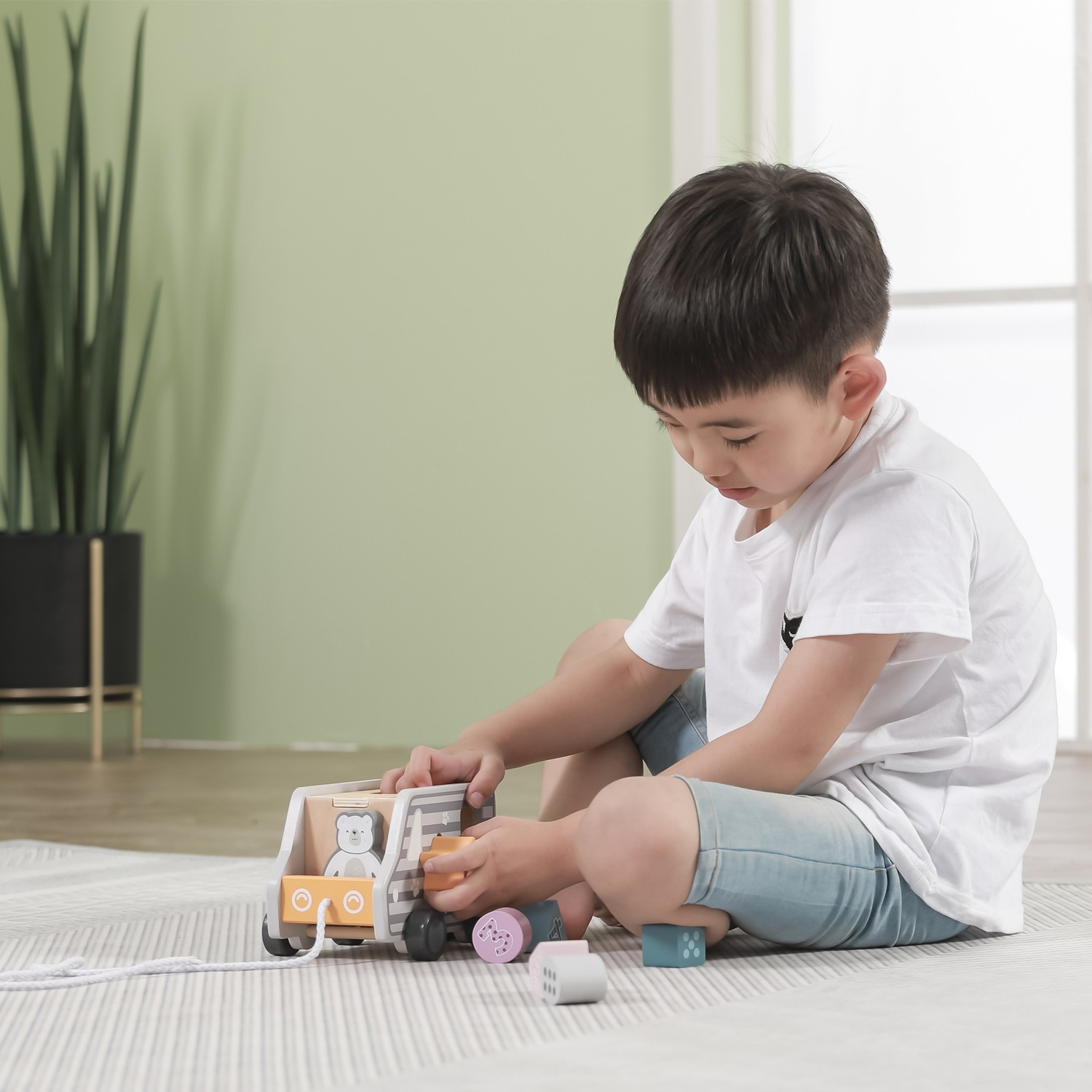 Criança a brincar com brinquedo de madeira num tapete cinzento claro numa sala com parede verde clara e planta