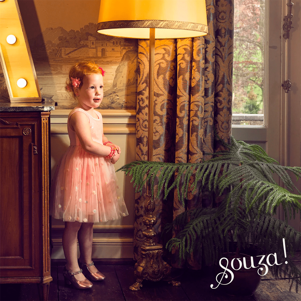 Menina com vestido rosa claro e sapatos dourados ao lado de candeeiro antigo e cortinas decorativas.