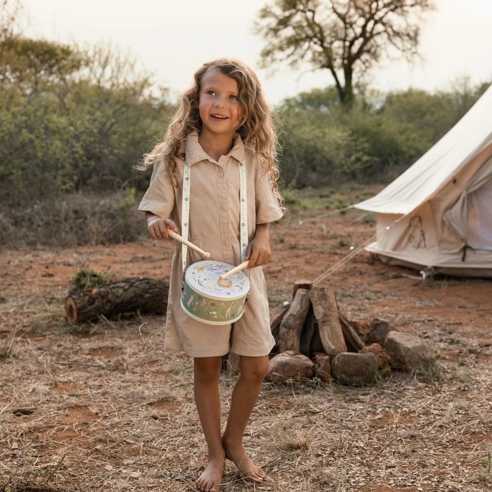 Menina com vestido bege a tocar tambor verde decorado, ao ar livre perto de tenda branca