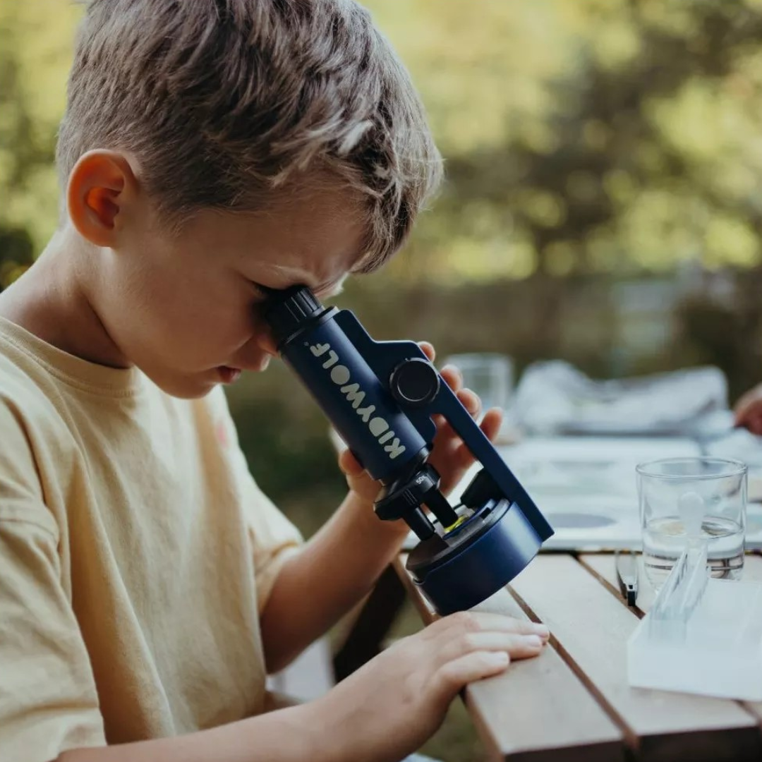 Criança olhando por um microscópio azul escuro com texto KID WOK ao ar livre