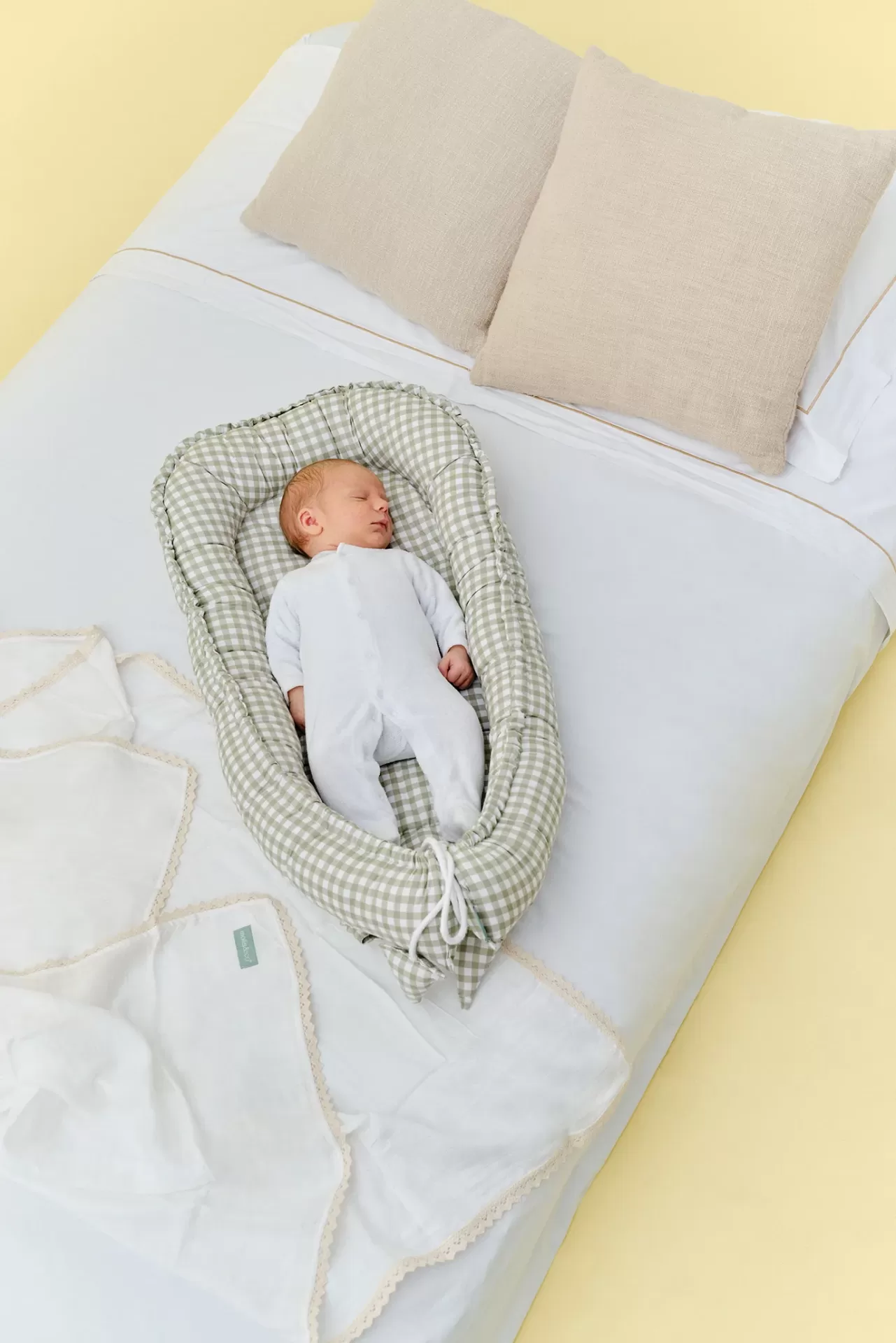 Bebé a dormir num ninho almofadado xadrez verde e branco sobre cama branca com almofadas bege.