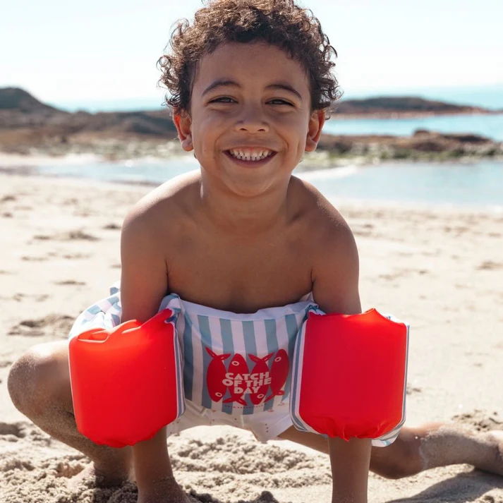 Criança na praia com colete flutuador azul e branco e braços infláveis vermelhos com texto e peixes