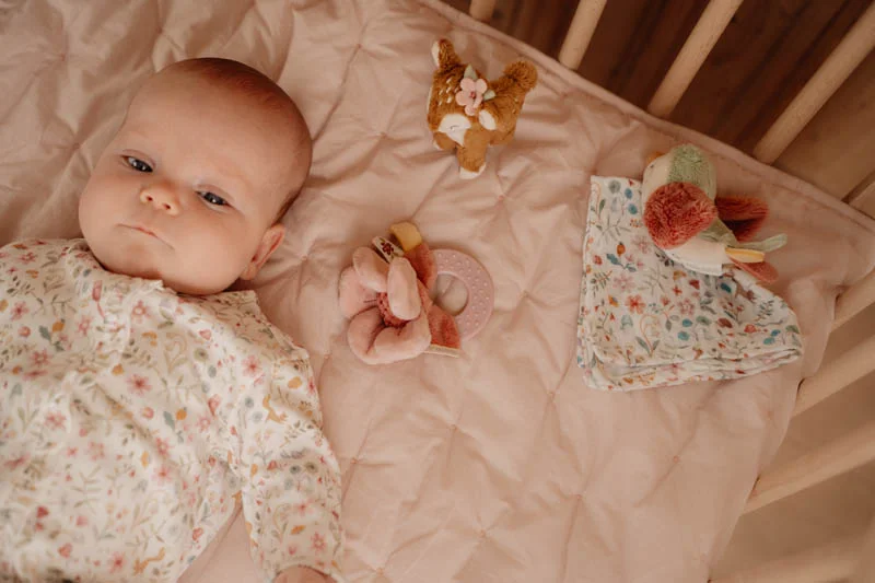 Bebé deitado em colchão cor de rosa num berço com brinquedos macios e manta floral