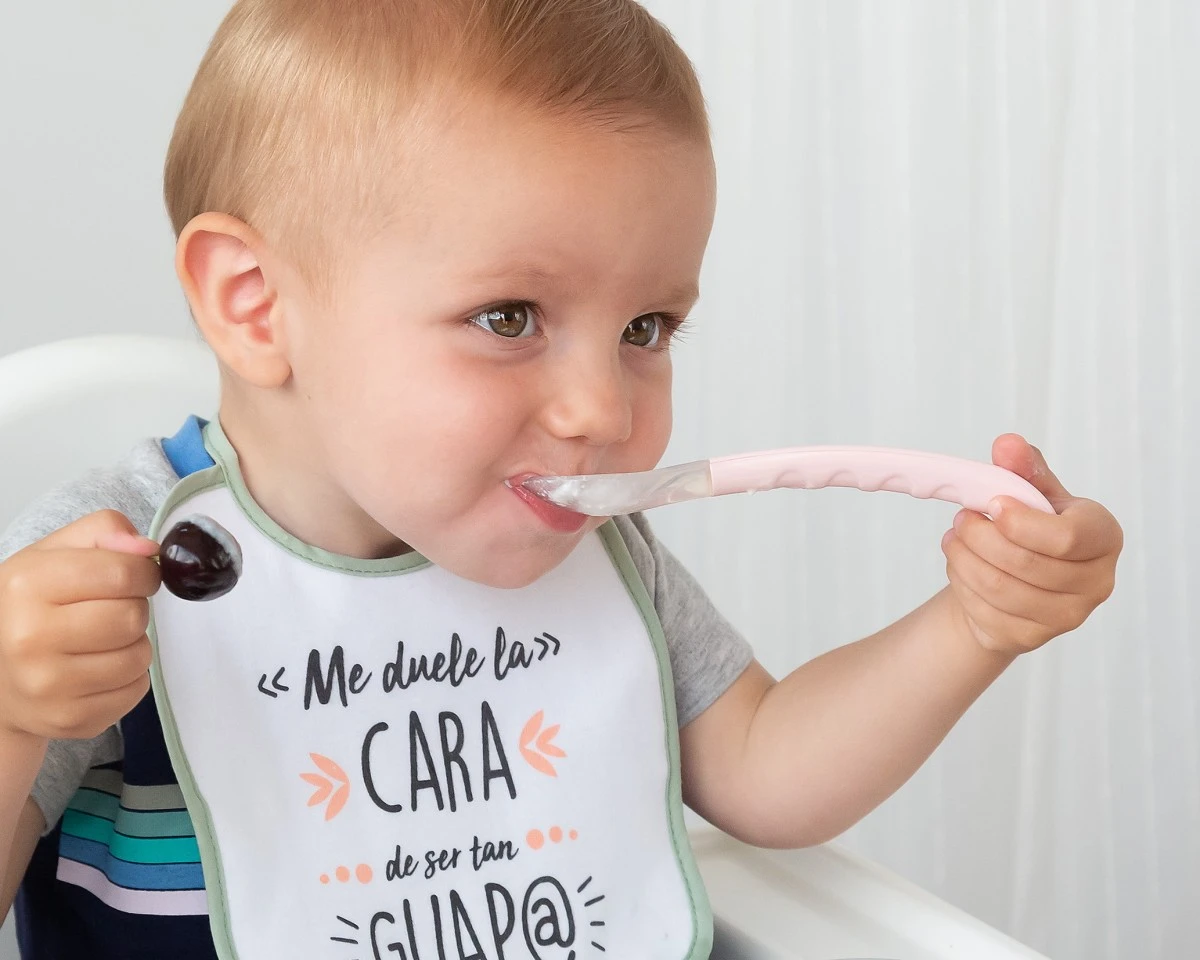 Bebé com babete branco com texto a ser alimentado com colher rosa.
