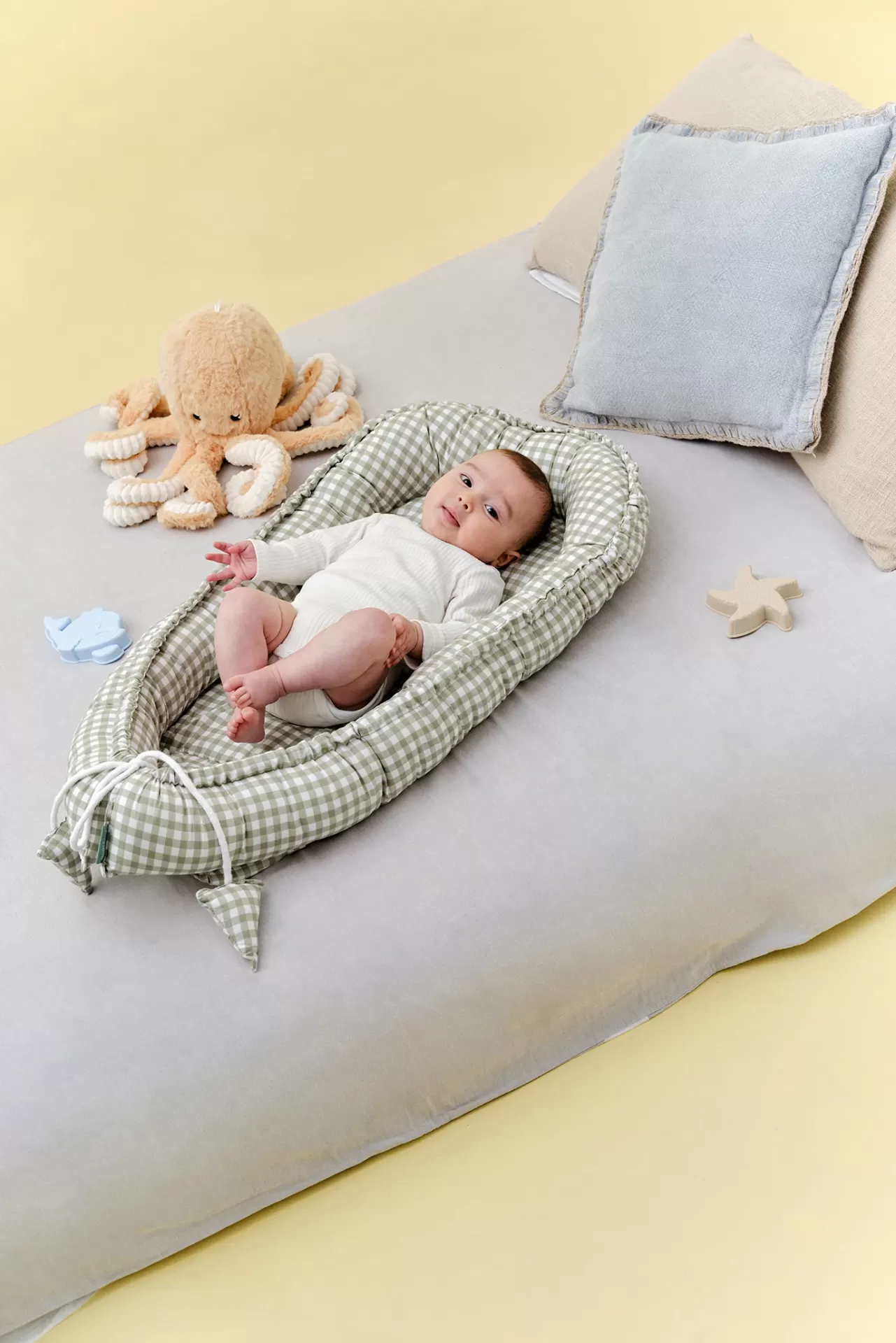 Berço portátil acolchoado xadrez verde claro com bebé deitado em almofada cinza e brinquedos por perto