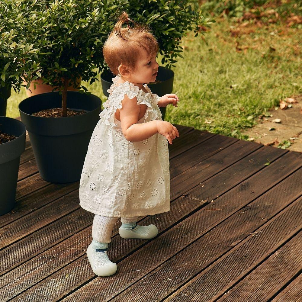 Bebé com vestido branco e sapatos azuis claros em madeira exterior.