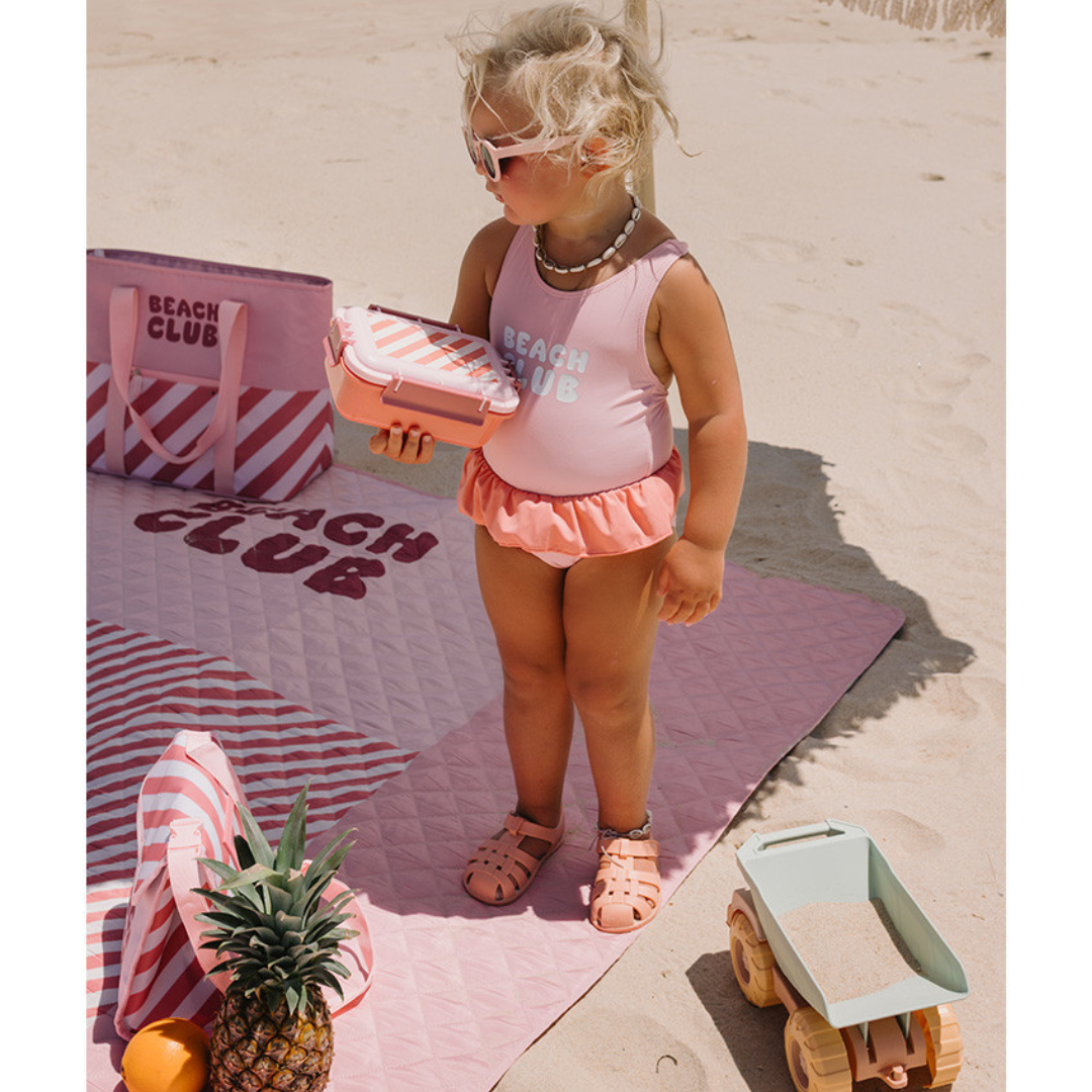 Criança pequena com fato de banho rosa, manta e saco de praia cor-de-rosa com texto BEÁCH CLUB, óculos de sol e brinquedo de carrinho na areia