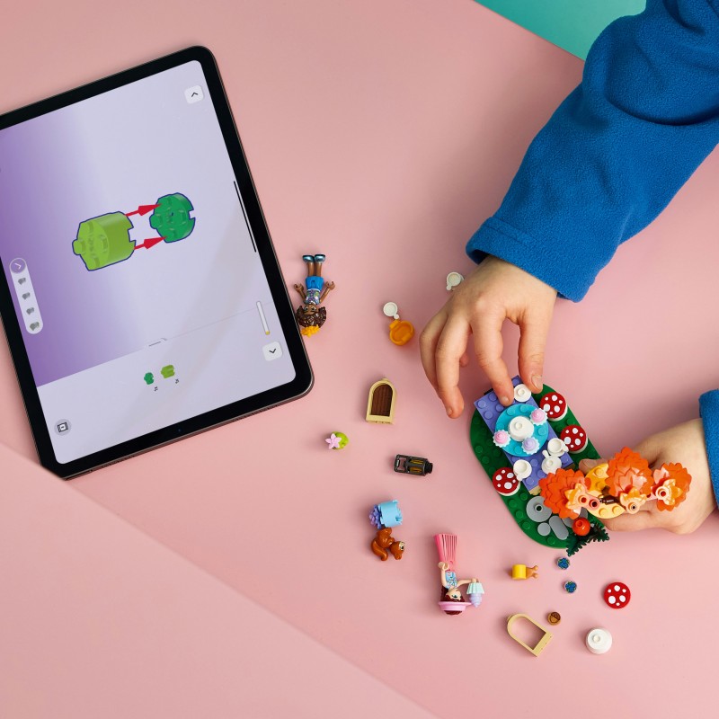 Criança a montar brinquedo de blocos coloridos junto a tablet com instruções numa superfície rosa.