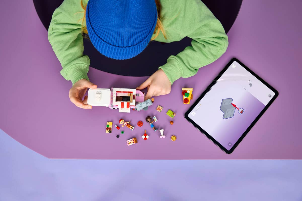 Pessoa a montar brinquedo de blocos de construção coloridos sobre mesa roxa com miniaturas e tablet preto.