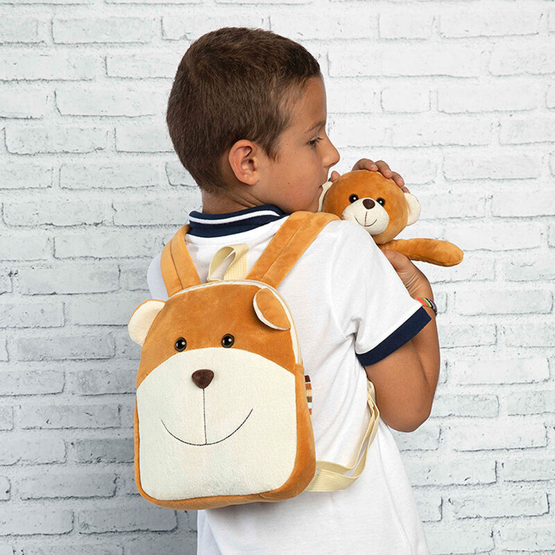 Mochila em forma de urso castanho e peluche castanho nas mãos de menino junto a parede branca de tijolos.