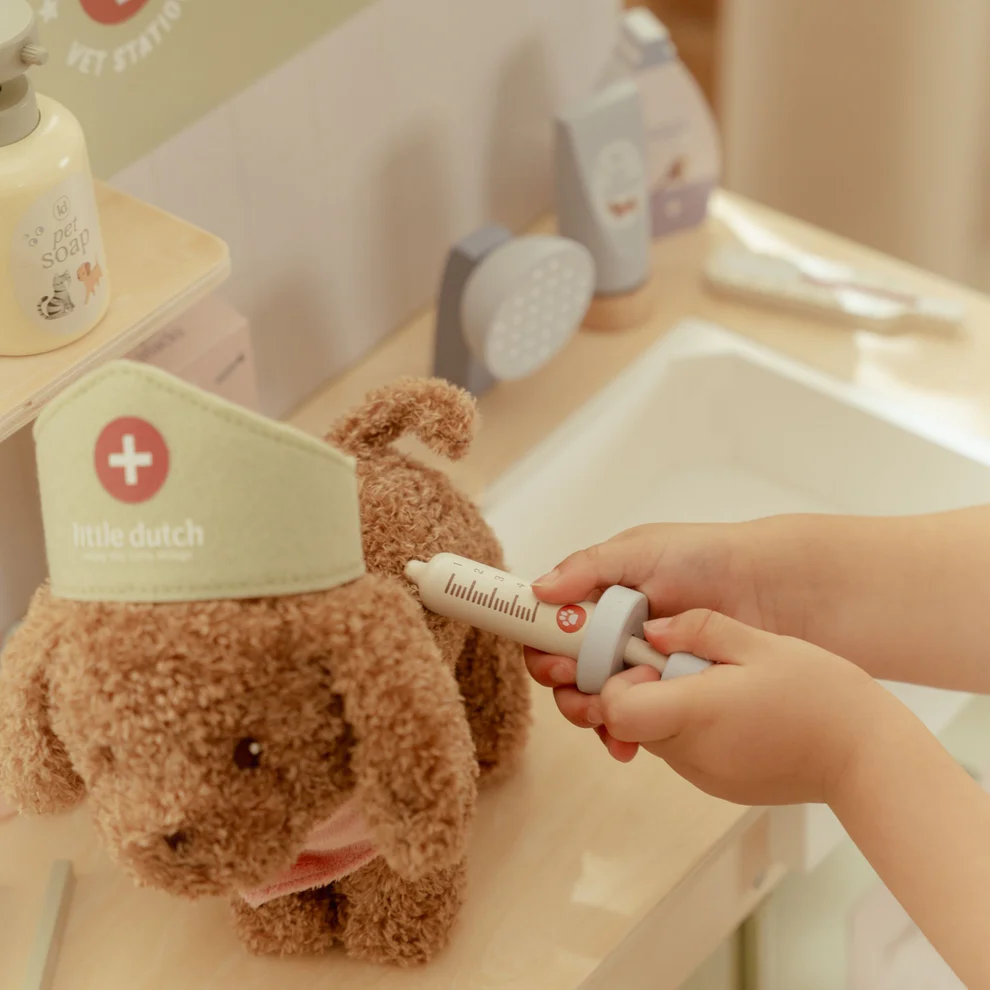 Brinquedo de cão de pelúcia com chapéu little dutch e uma mão segurando seringa de brinquedo