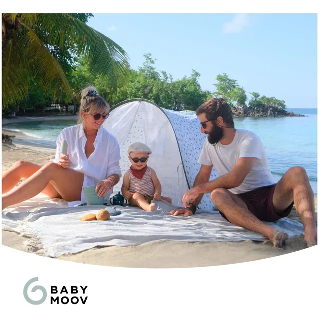 Familia numa praia com uma tenda de bebé branca com padrão de folhas, sentados num tapete cinzento